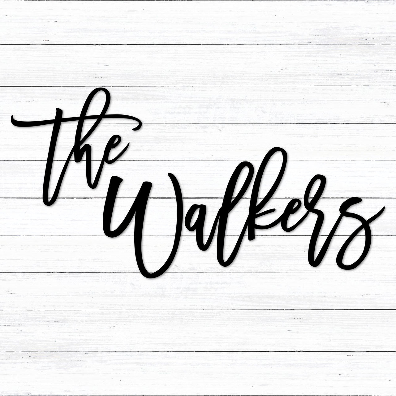Custom black powder coated last name the Walkers metal sign in an elegant and fun handwritten script font placed on a white washed shiplap wall. The personalized sign is in two different pieces: the and Walkers. 